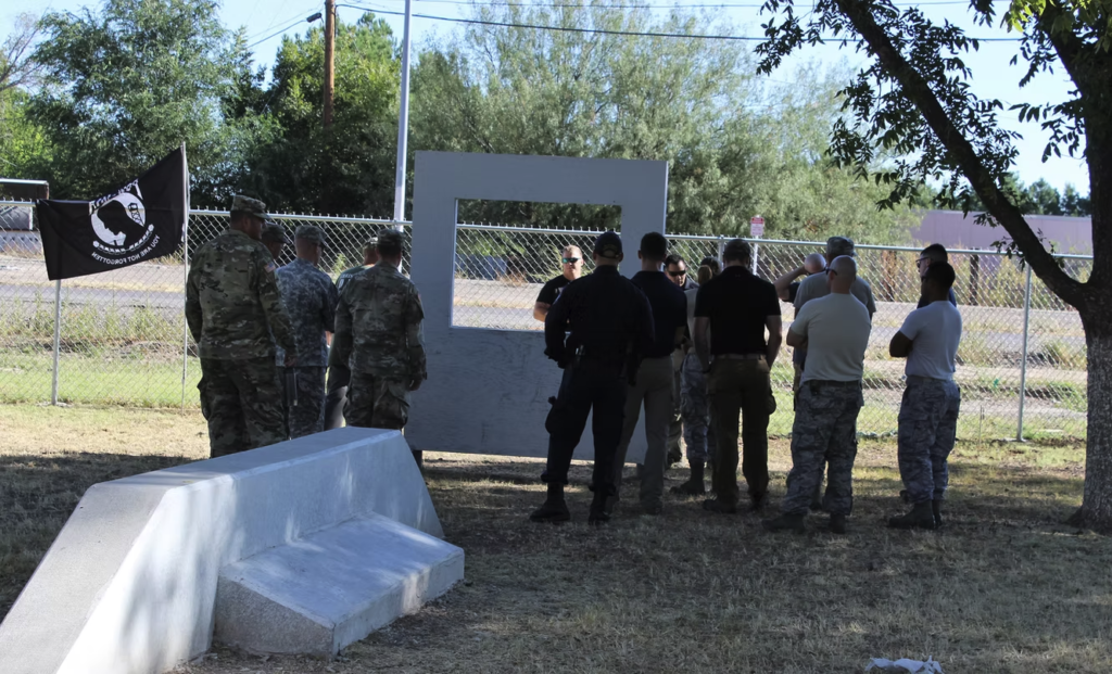 Service members and law enforcement officers gathered at a K9 training or memorial event beside a POW/MIA flag